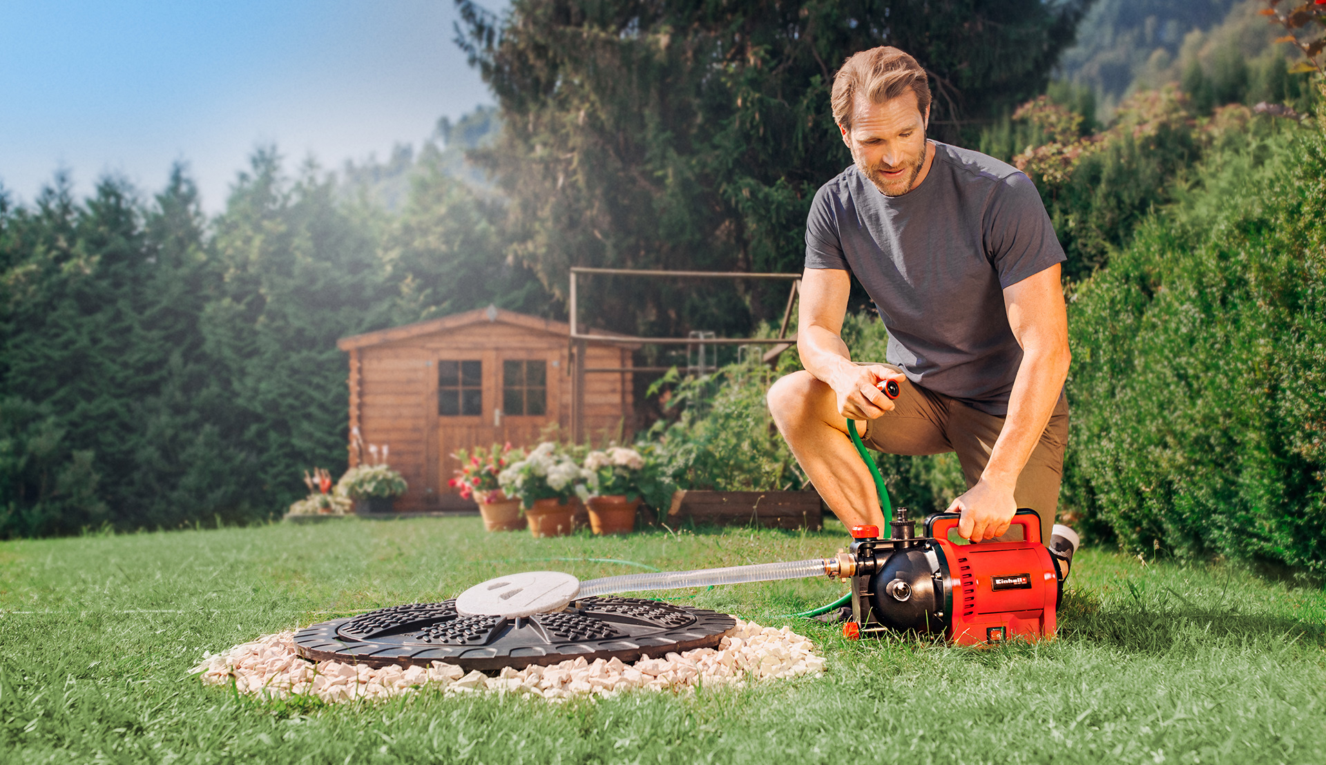 A man kneels in the garden in front of an Einhell garden pump connected to a cistern.