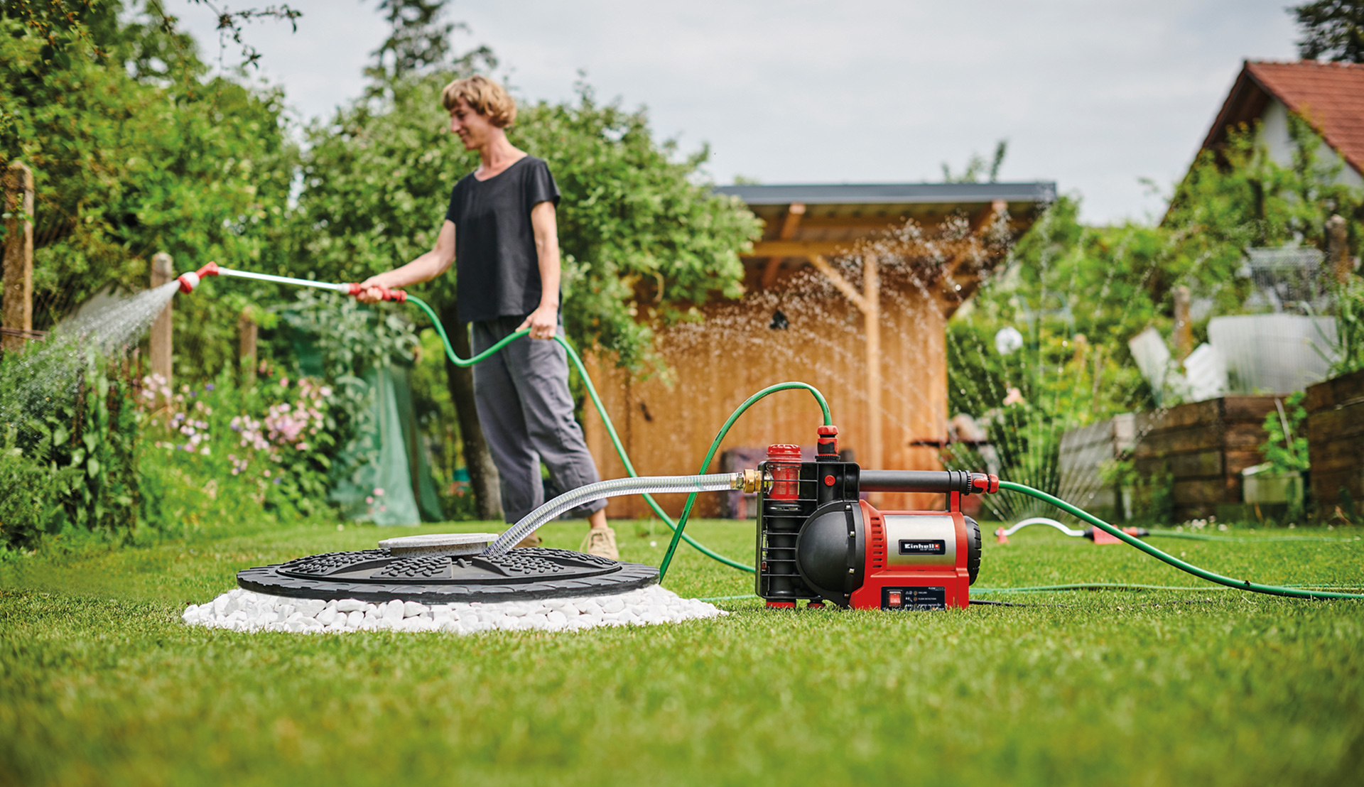 A woman waters her flowers and shrubs with the help of an Einhell garden pump, which supplies water to an irrigation sprinkler and a lawn sprinkler.