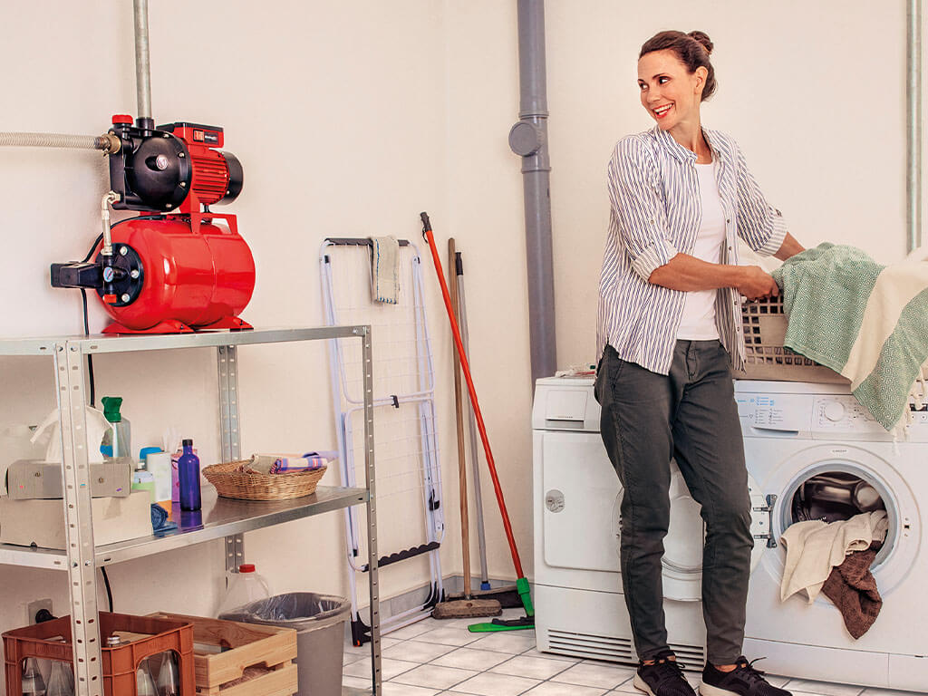 A woman stands in front of the washing machine with her laundry basket and looks at the connected Einhell domestic waterworks.