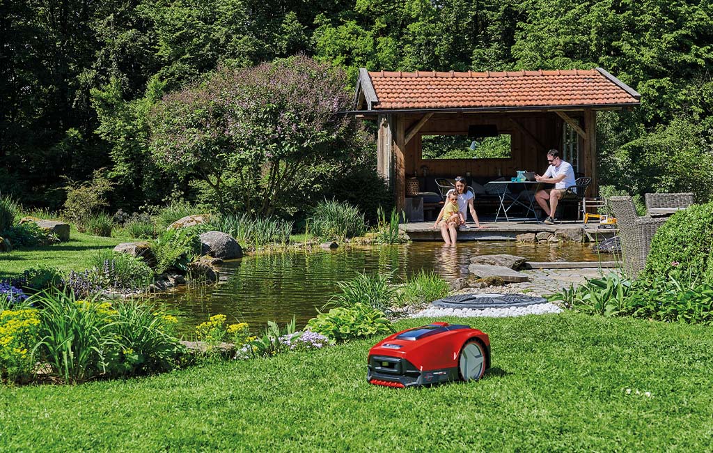 A robotic mower cutting the lawn in a garden, with a pond in the background.