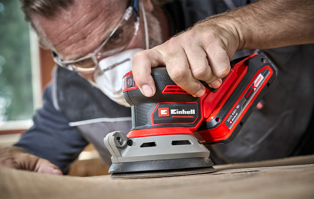 Man with protective mask and goggles sanding wood with an Einhell Professional cordless rotating sander