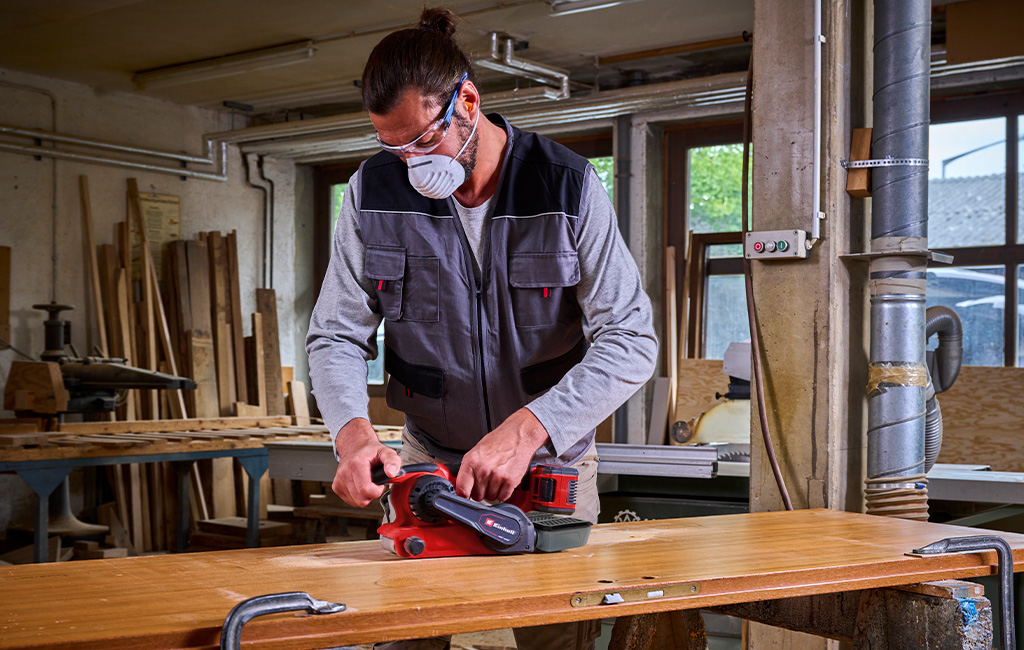 Man in a workshop sanding a wooden door using an Einhell Professional cordless belt sander