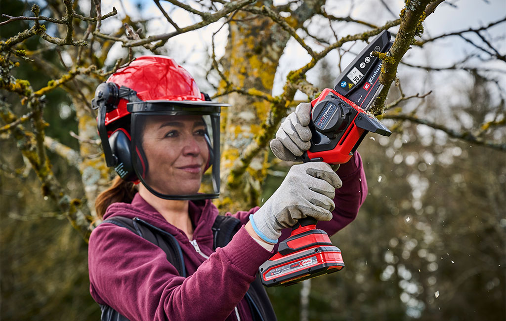 Woman wearing a forestry helmet sawing off the branch of a tree with a cordless pruning chainsaw.