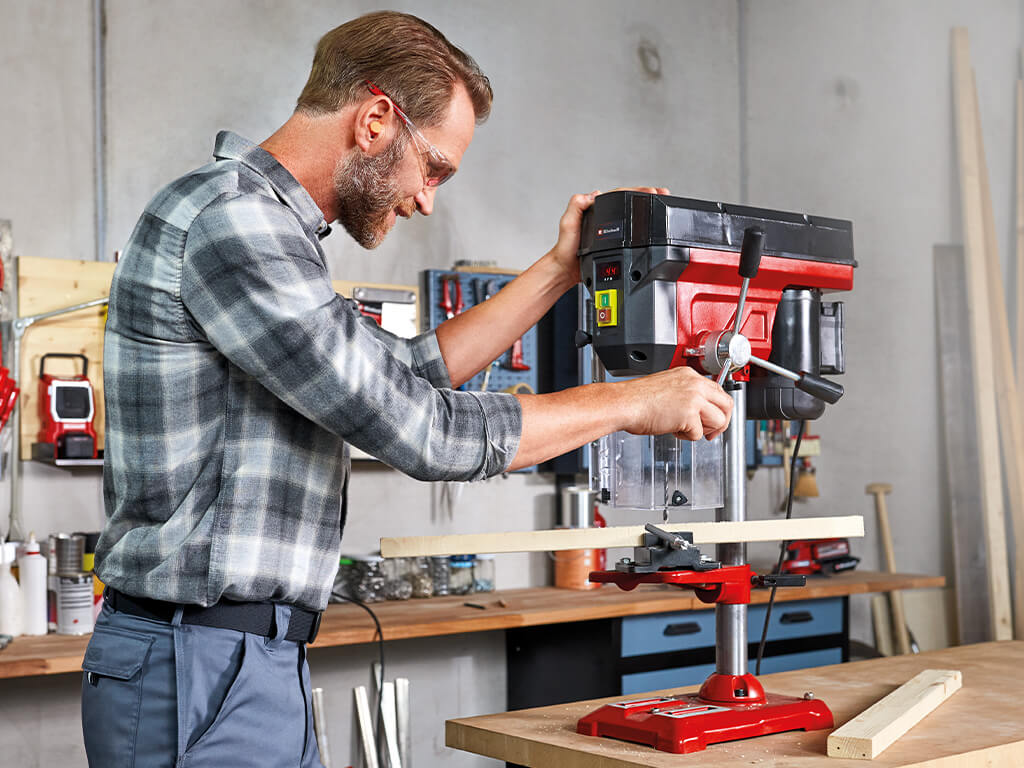 Man moves the turnstile of a bench drill to drill a hole in a wooden slat.