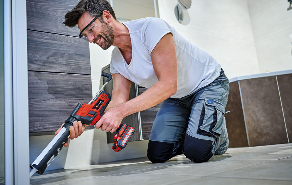 A man kneeling on the floor, working with a cordless sealing gun.