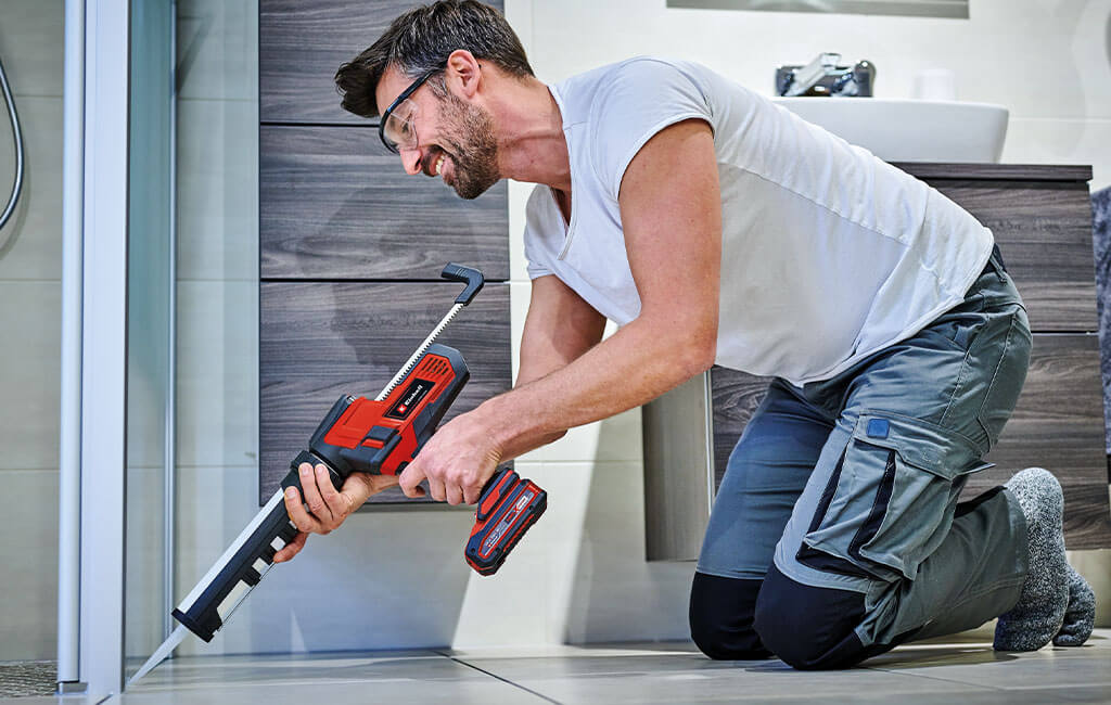 A man kneeling on the floor in the bathroom and applying silicone as a sealant between the shower and the floor.