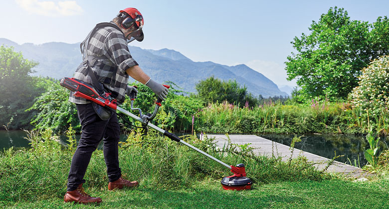 a man mows the lawn in front of a footbridge with a battery-powered scythe