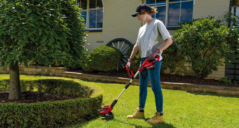 a woman trims the lawn along a bush that is around a tree